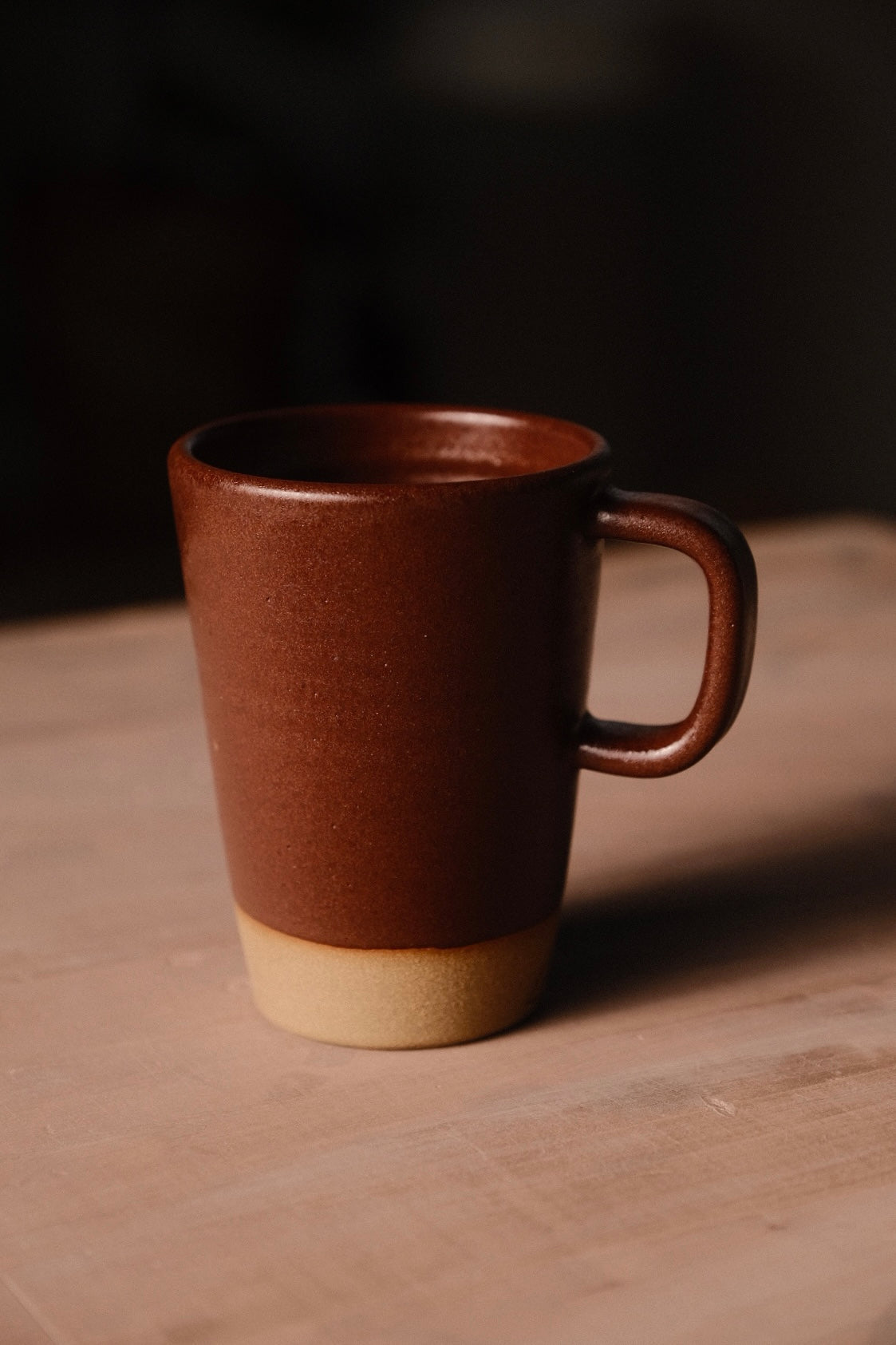 Reddish-brown Settle Ceramics Wander Latte Mug on a wooden tabletop, viewed slightly from above, showing its tall tapered ceramic body, rounded rectangular handle, glossy interior and contrasting sandy-beige base.