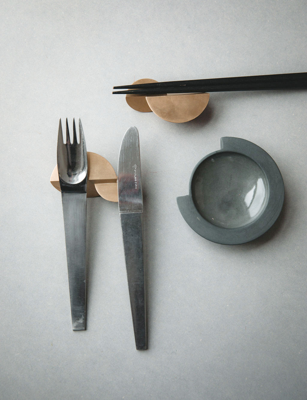 Empty graphite Tortuga Forma Kaolin Sauce Dish beside a fork, knife and black chopsticks on brass rests, showing its glossy circular well and notched rim on a grey tabletop.