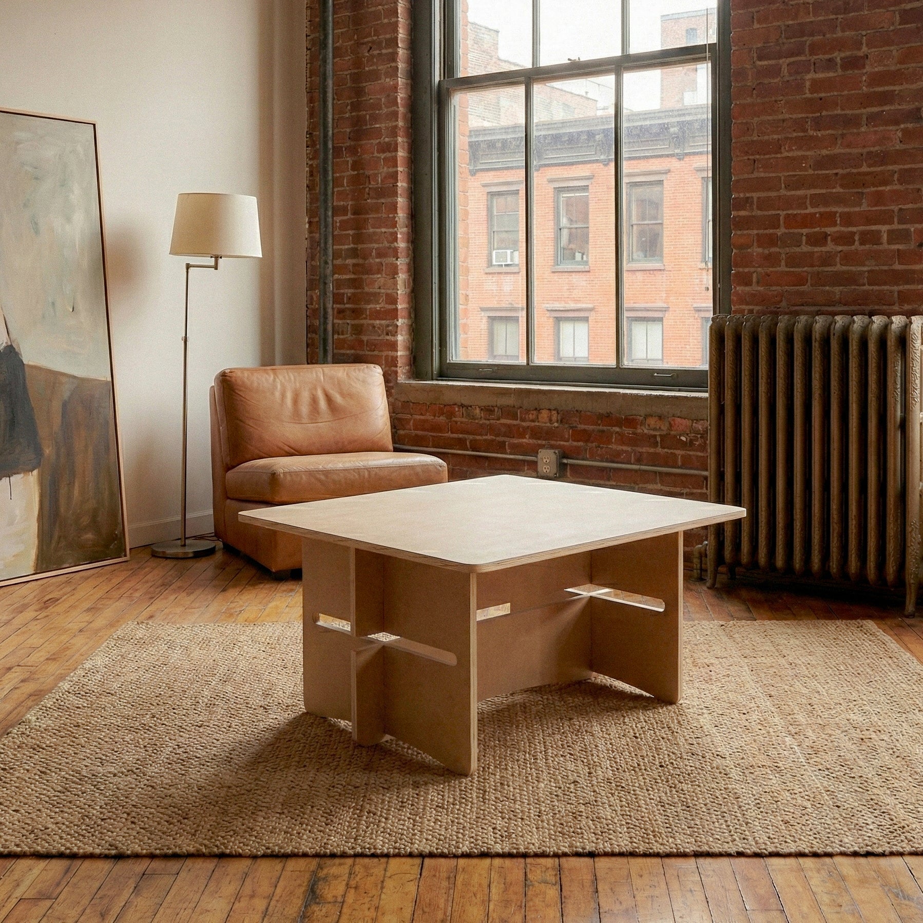 Williamsburg wood coffee table centered on a woven rug in a loft living room with a leather chair, exposed brick and large windows