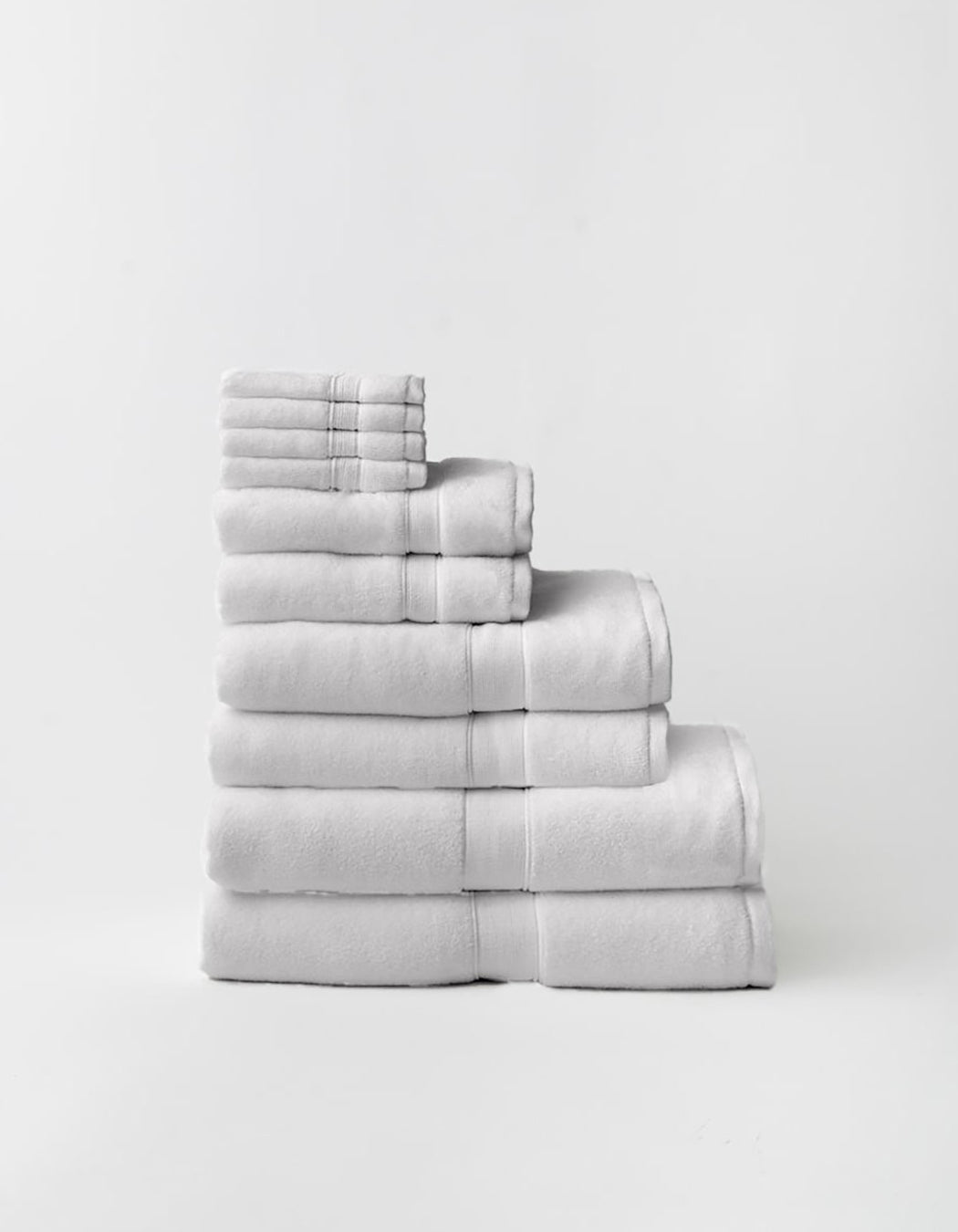 Light gray towels stacked in a stepped arrangement on a pale background, showing multiple sizes, thick folded edges, and woven border bands.
