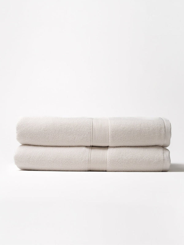 Two ivory Luxe Bath Sheets folded and stacked on a pale background, showing plush looped texture, thick folded edges, and matching woven border bands.