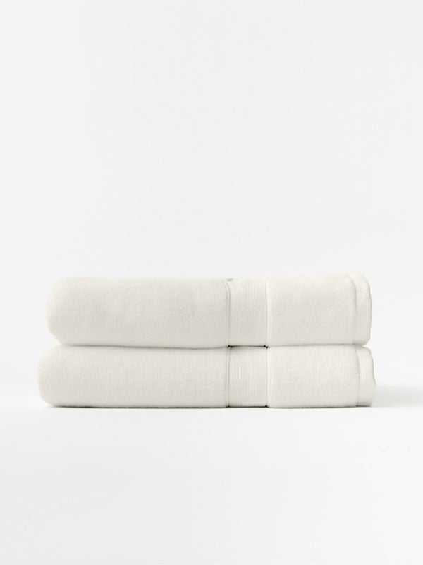 Two ivory bath towels folded and stacked on a white background, showing plush texture, thick folded edges, and matching woven border bands.