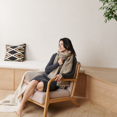 Woman seated in a wooden lounge chair with the neutral scarf wrapped warmly around her neck.
