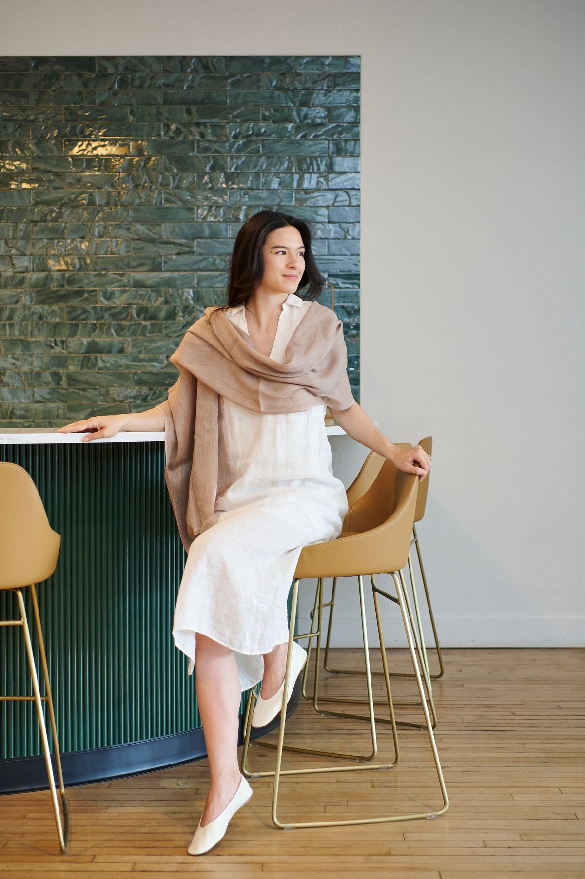 Woman seated at a modern counter wearing the Variously Jute Golden Brown linen-silk scarf loosely draped around her shoulders