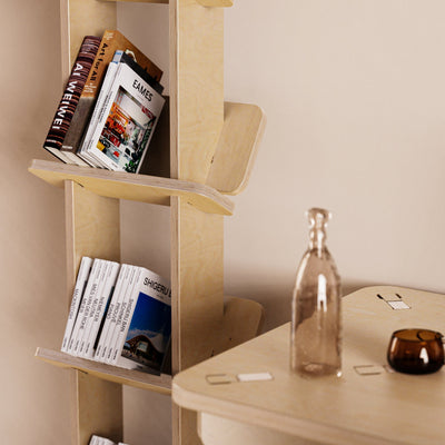Close-up of books and magazines resting on the Yankel bookcase’s angled natural wood shelves beside a tabletop