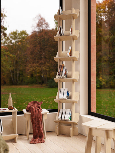 Yankel bookcase filled with books beside a large window overlooking autumn trees, with a rust-colored throw and stool nearby