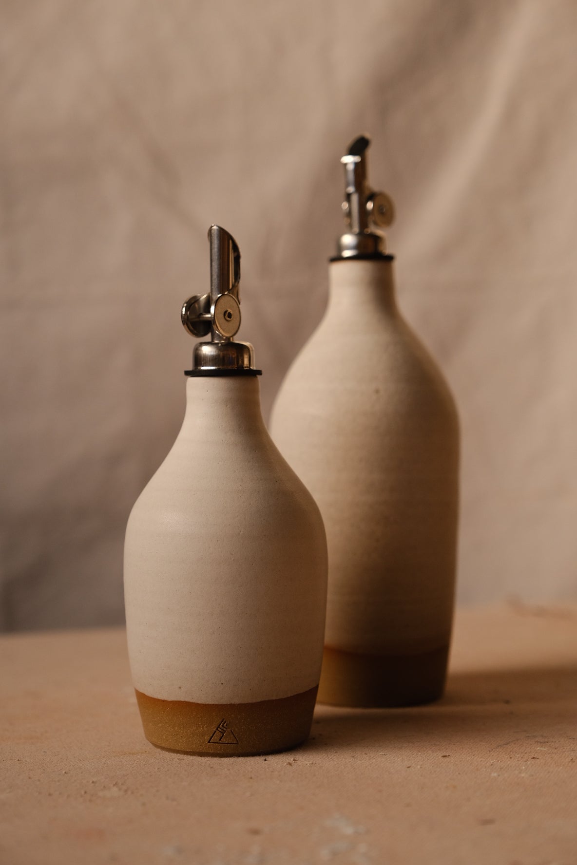Warm beige Settle Ceramics Wander Tall Oil Bottle beside a shorter matching bottle, showing rounded ceramic bodies, fine surface speckles and metal pouring spouts on a tan tabletop.
