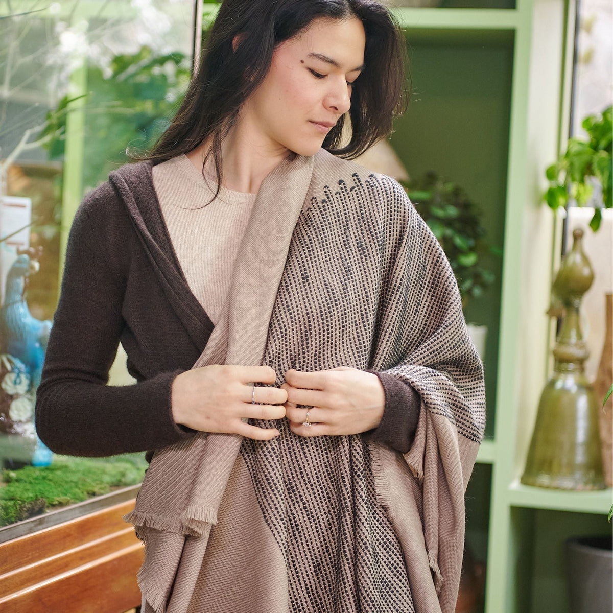 Woman wrapped in the Flo Brown merino throw in a plant-filled room, highlighting its soft drape and textured design