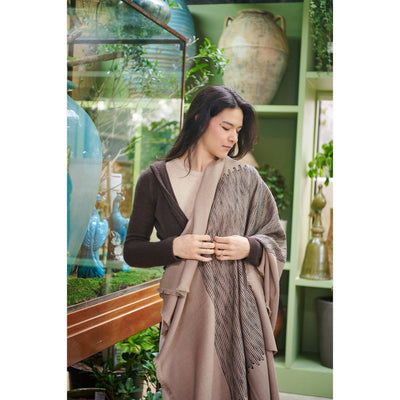 Full-length view of a woman wrapped in the Flo Brown merino wool throw beside a sunlit window and indoor plants