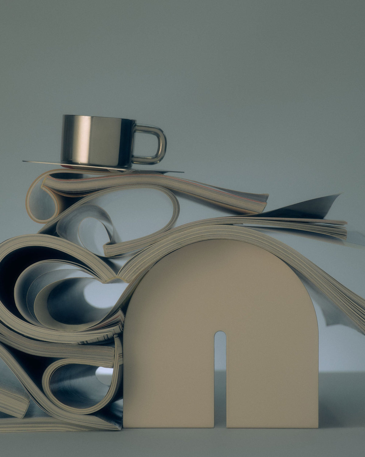 Beige bookend from Tortuga Forma Dumbo Bookend Pairs - The Architect Collection beside loosely rolled and stacked magazines, with a metal cup above, against a muted blue-green background.