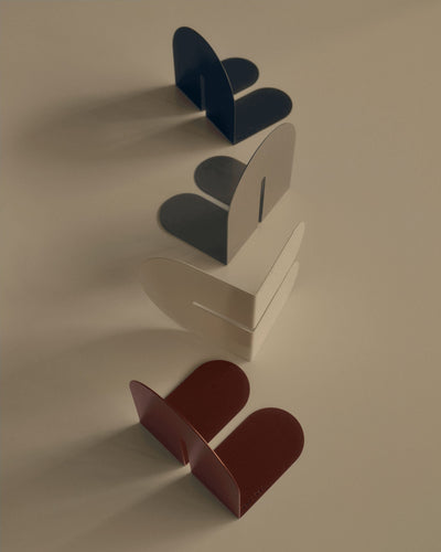 Tortuga Forma Dumbo Bookend Pairs - The Architect Collection in dark blue, taupe, cream and reddish brown arranged diagonally on a beige surface, casting long shadows that emphasize their arched panels and projecting feet.