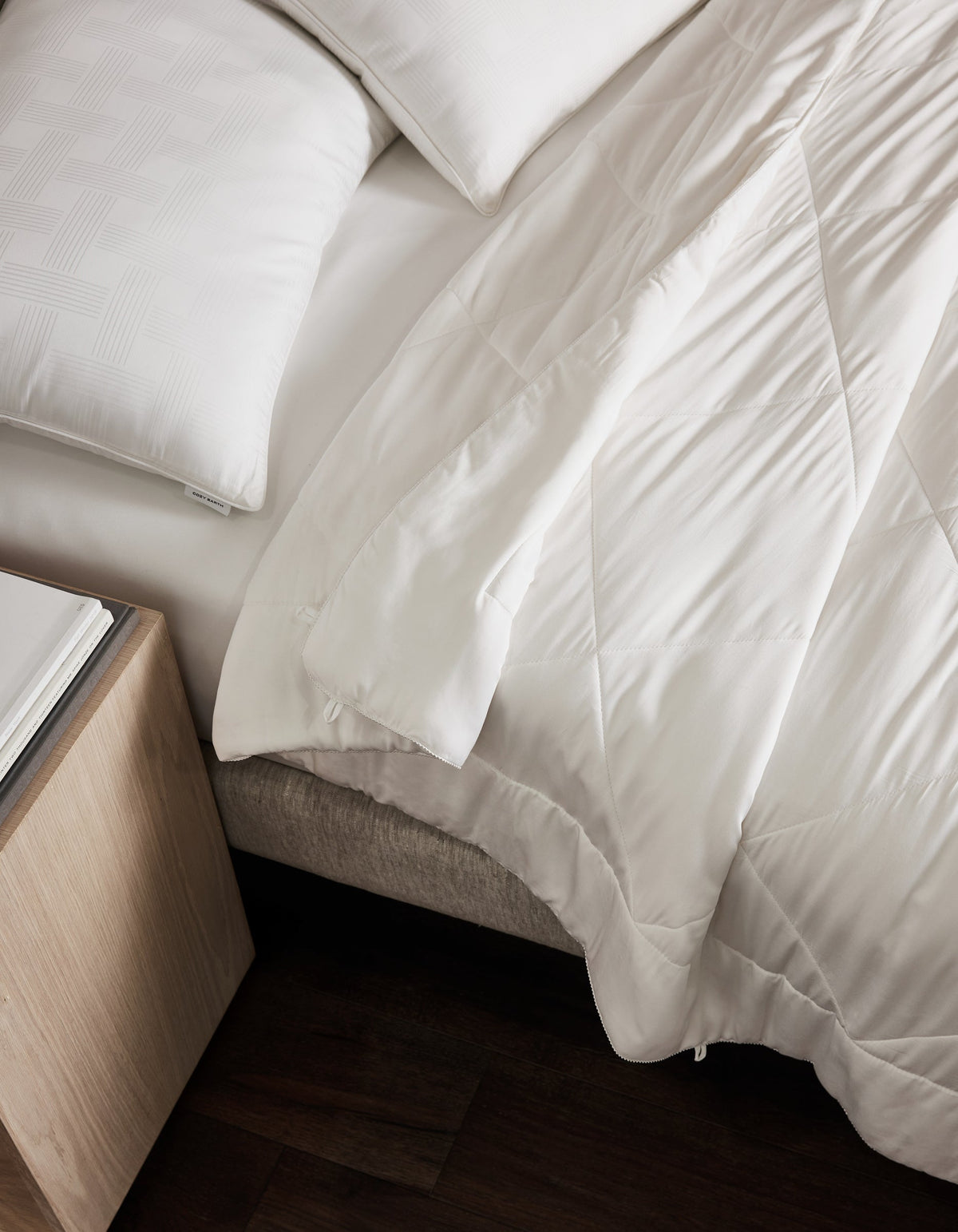Overhead angled view of the white comforter draped across a bed beside a wooden nightstand, showing broad quilted sections and soft folds.