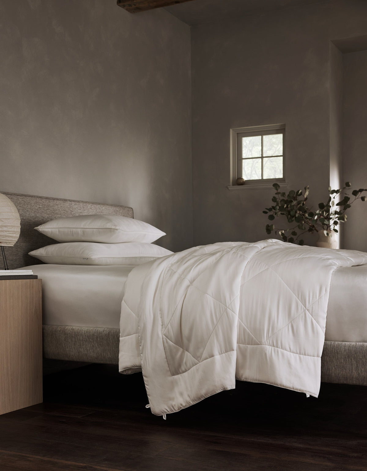 White comforter draped over the foot of a bed in a neutral bedroom, with stacked pillows, a wooden nightstand, and soft window light.