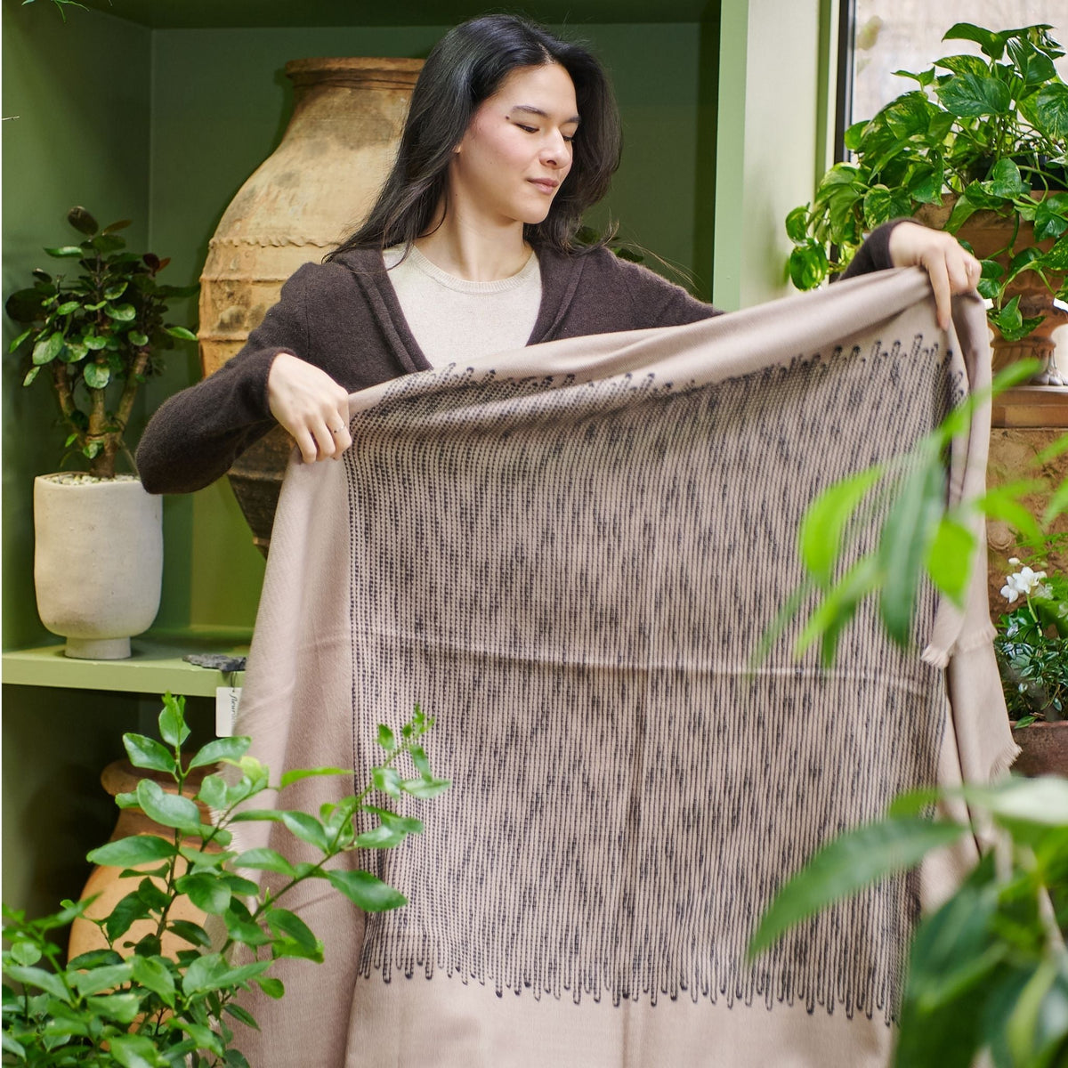 Woman holding the Variously Flo Brown merino wool throw open among green plants, displaying its taupe border and dark textured center panel