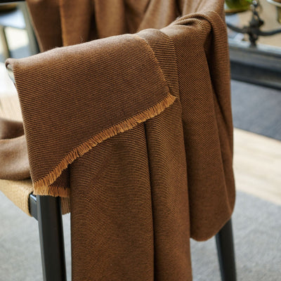 Hay Brown handwoven throw folded over a chair arm, highlighting its rich brown tone and contrasting golden fringe