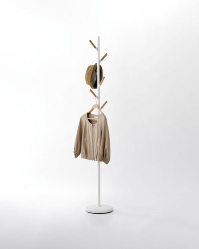 White Yamazaki Home Coat Rack (70" H) - Steel on a pale background, with a slim upright pole, round base and staggered wood-tipped branches holding a tan hat and beige jacket.