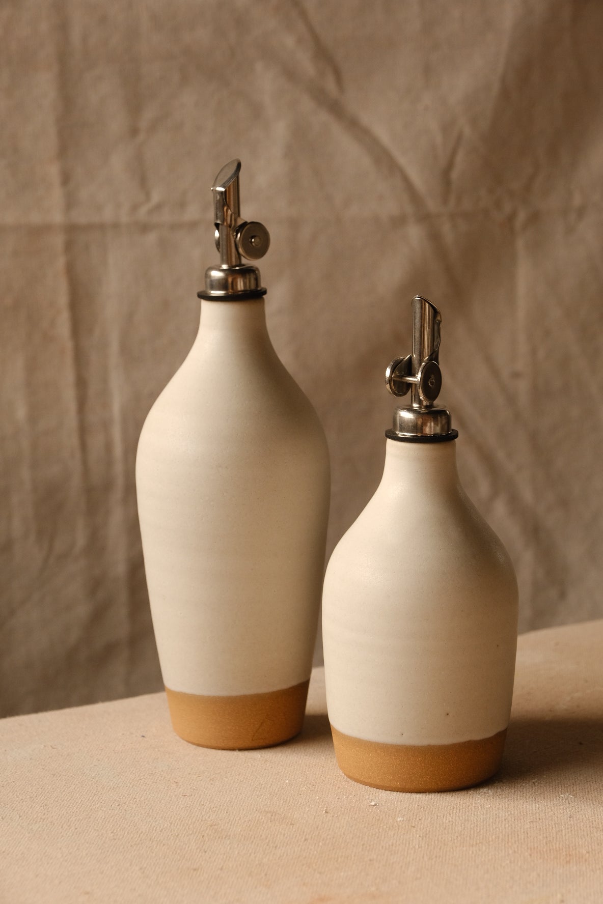 Ivory Settle Ceramics Wander Tall Oil Bottle behind a shorter matching bottle, with gently tapered bodies, narrow necks, metal pourers and golden-tan bands at the bases.