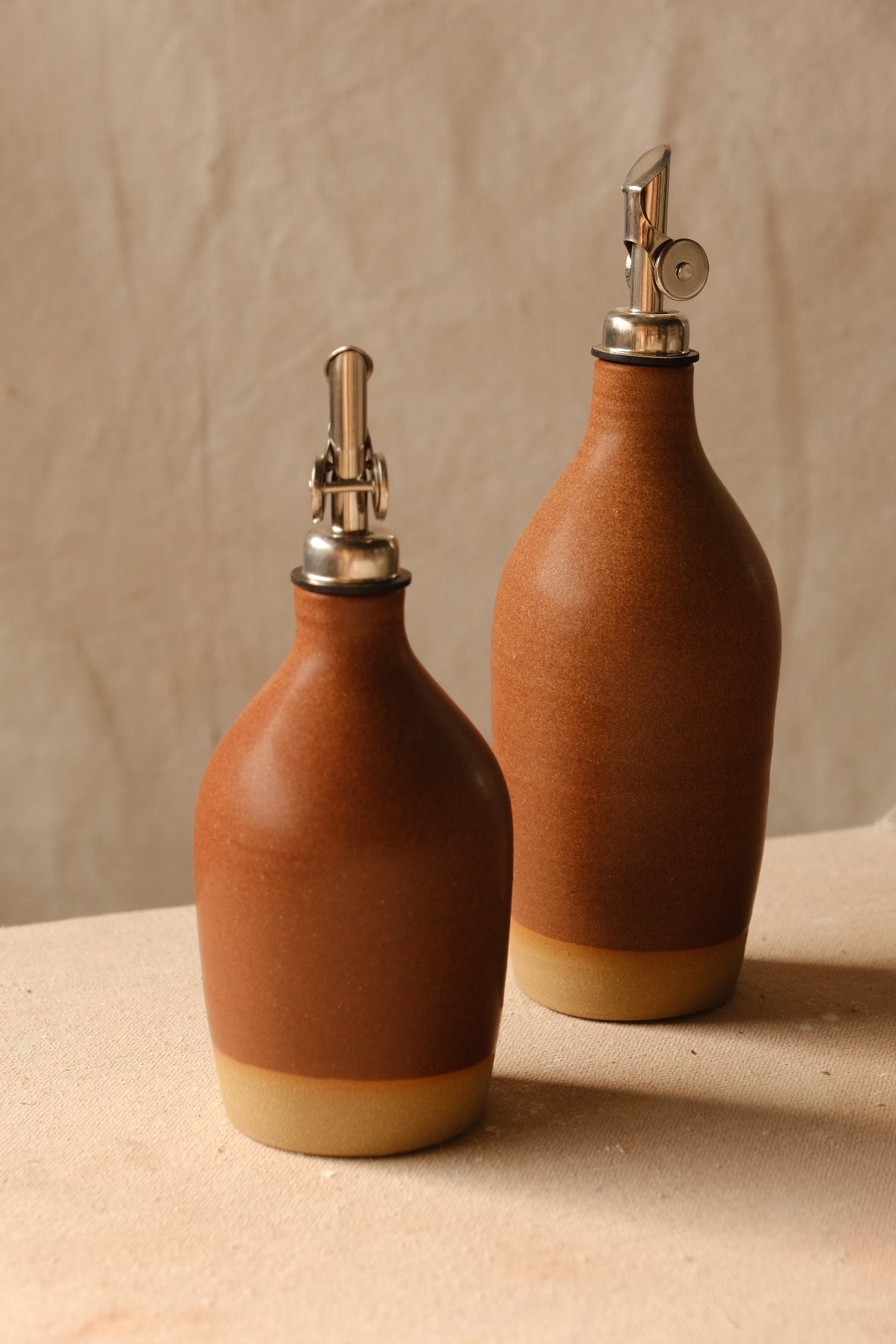 Rust-orange Settle Ceramics Wander Oil Bottle in front of a taller matching bottle against beige fabric, showing finely speckled ceramic bodies, metal pouring spouts and pale ochre bases.
