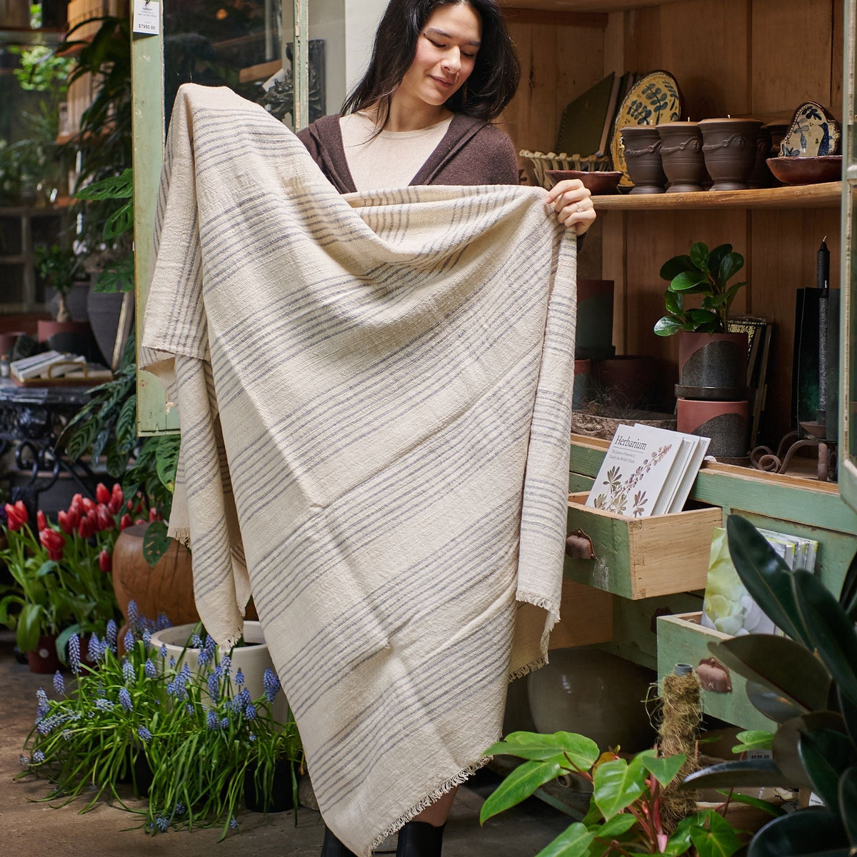 Woman holding the Silica Chalk off-white and gray striped throw open in a plant-filled artisan shop