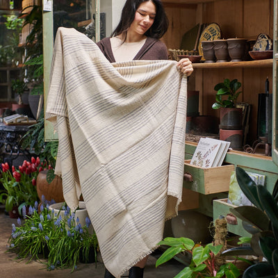 Woman holding the Silica Chalk off-white and gray striped throw open in a plant-filled artisan shop