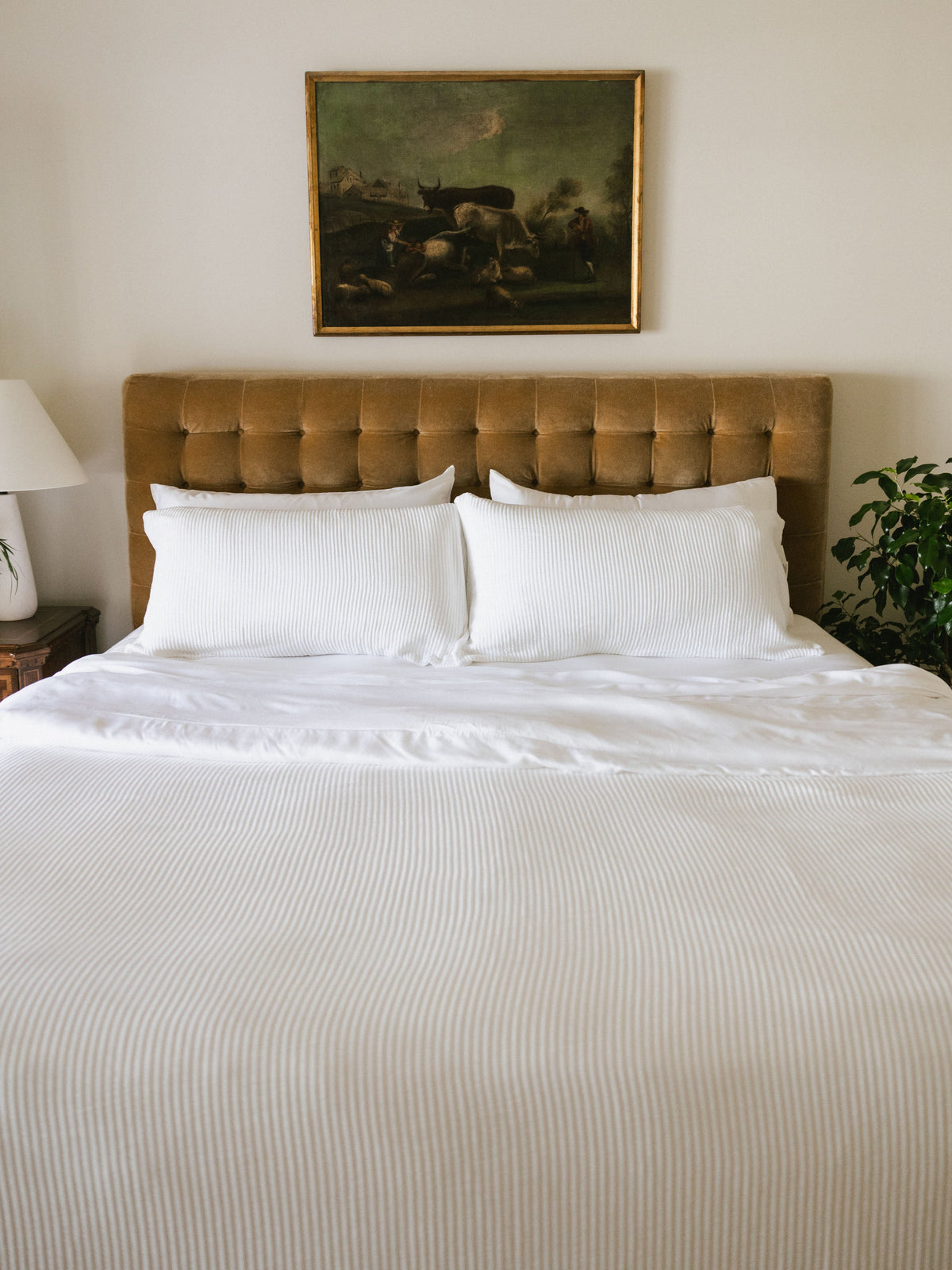 White ribbed coverlet spread across a bed with matching white pillows, a tan tufted headboard, and a framed painting above.
