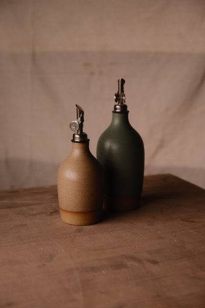 Deep green Settle Ceramics Wander Tall Oil Bottle behind a shorter tan bottle, with metal pourers catching the light against a draped beige backdrop.