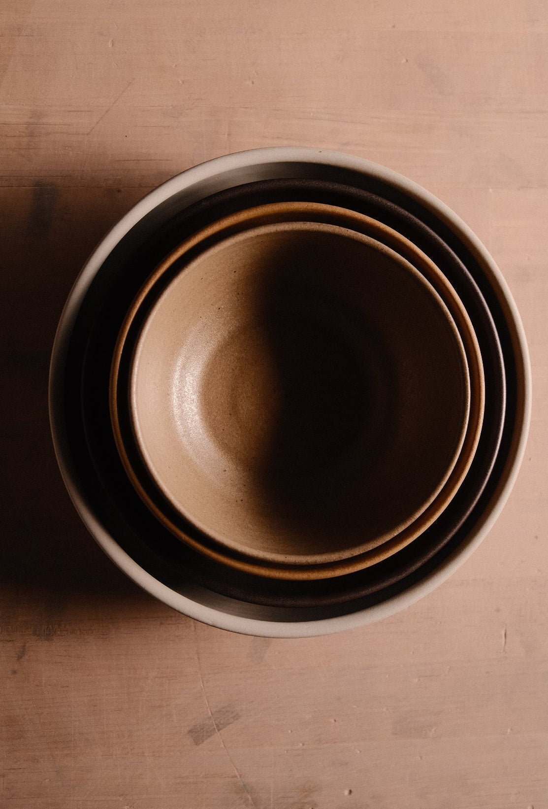 Overhead view of nested Settle Ceramics Wander Soup Bowls in brown and cream tones, forming concentric circles around the smallest bowl’s golden-brown interior on a wooden surface.