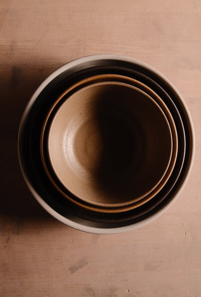 Overhead view of nested Settle Ceramics Wander Soup Bowls in brown and cream tones, forming concentric circles around the smallest bowl’s golden-brown interior on a wooden surface.