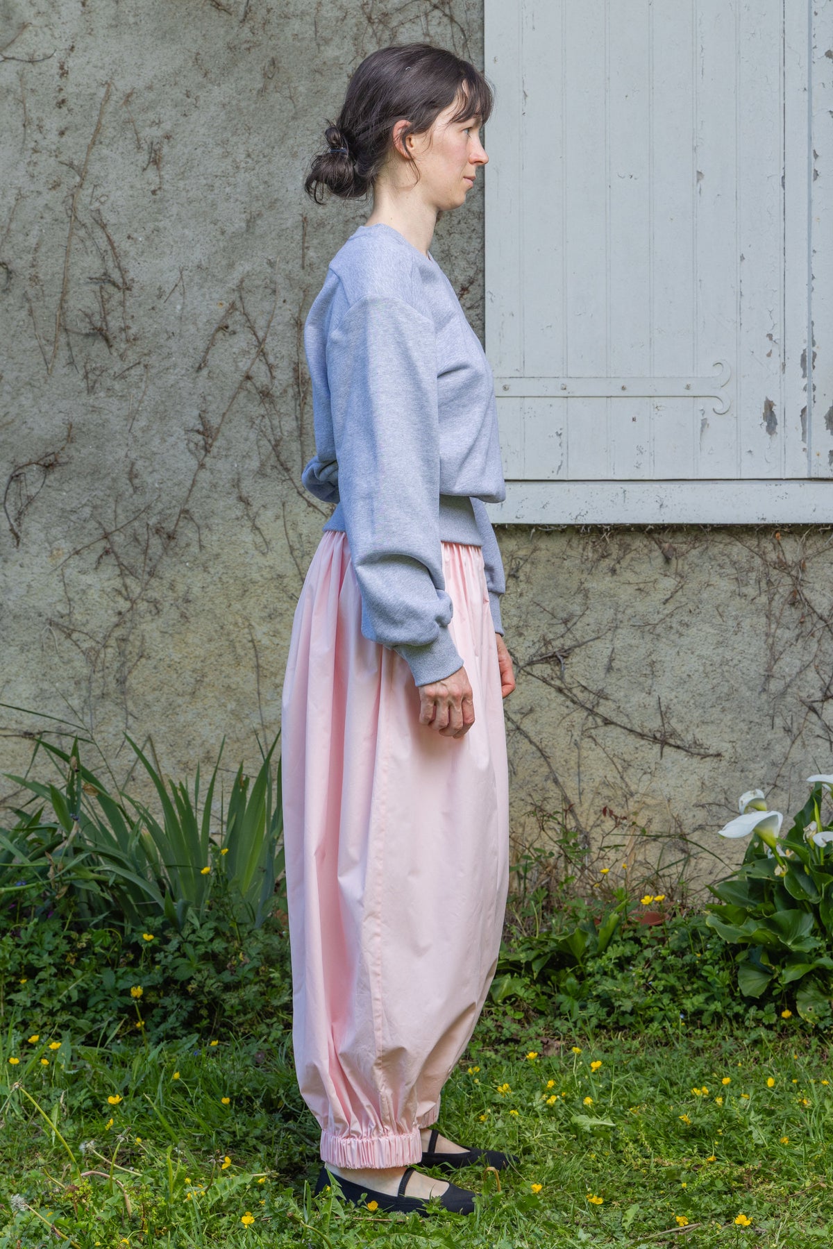Side view of a model wearing the light gray Paulette Madeleine Top with pale pink gathered trousers, showing its waist-length hem and roomy sleeve beside a stone wall and pale shutter.
