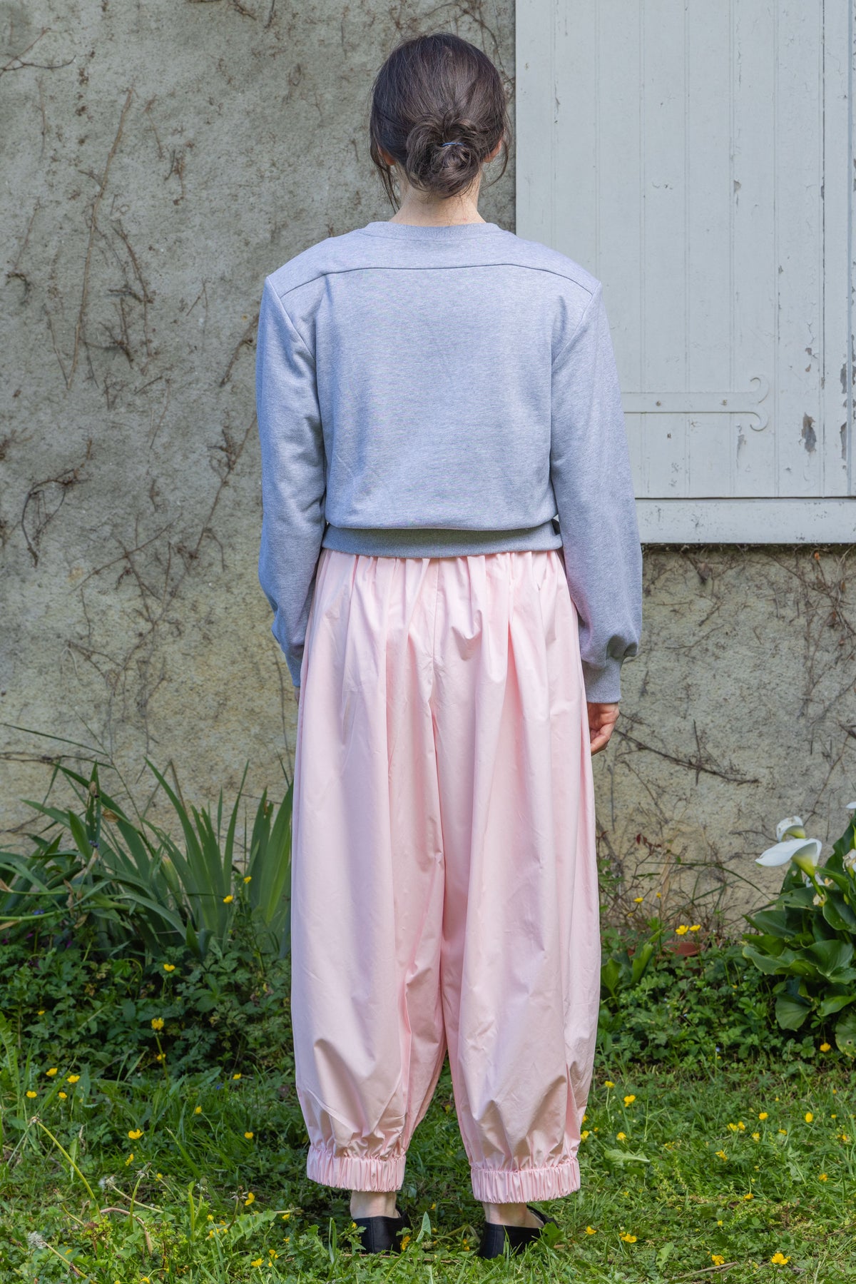 Back view of a model wearing the light gray Paulette Madeleine Top with pale pink trousers, showing the straight shoulder seams, plain back, and wide waistband.