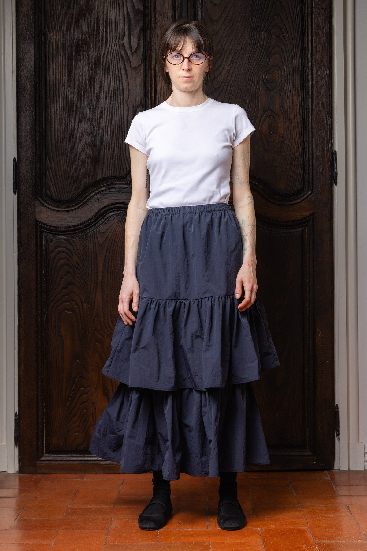 Model wearing the dark navy Paulette Louise Tier Skirt with a tucked-in white T-shirt and black ankle boots, standing before dark wooden doors and showing the gathered waist and two flowing lower tiers.