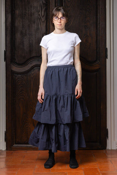 Model wearing the dark navy Paulette Louise Tier Skirt with a tucked-in white T-shirt and black ankle boots, standing before dark wooden doors and showing the gathered waist and two flowing lower tiers.