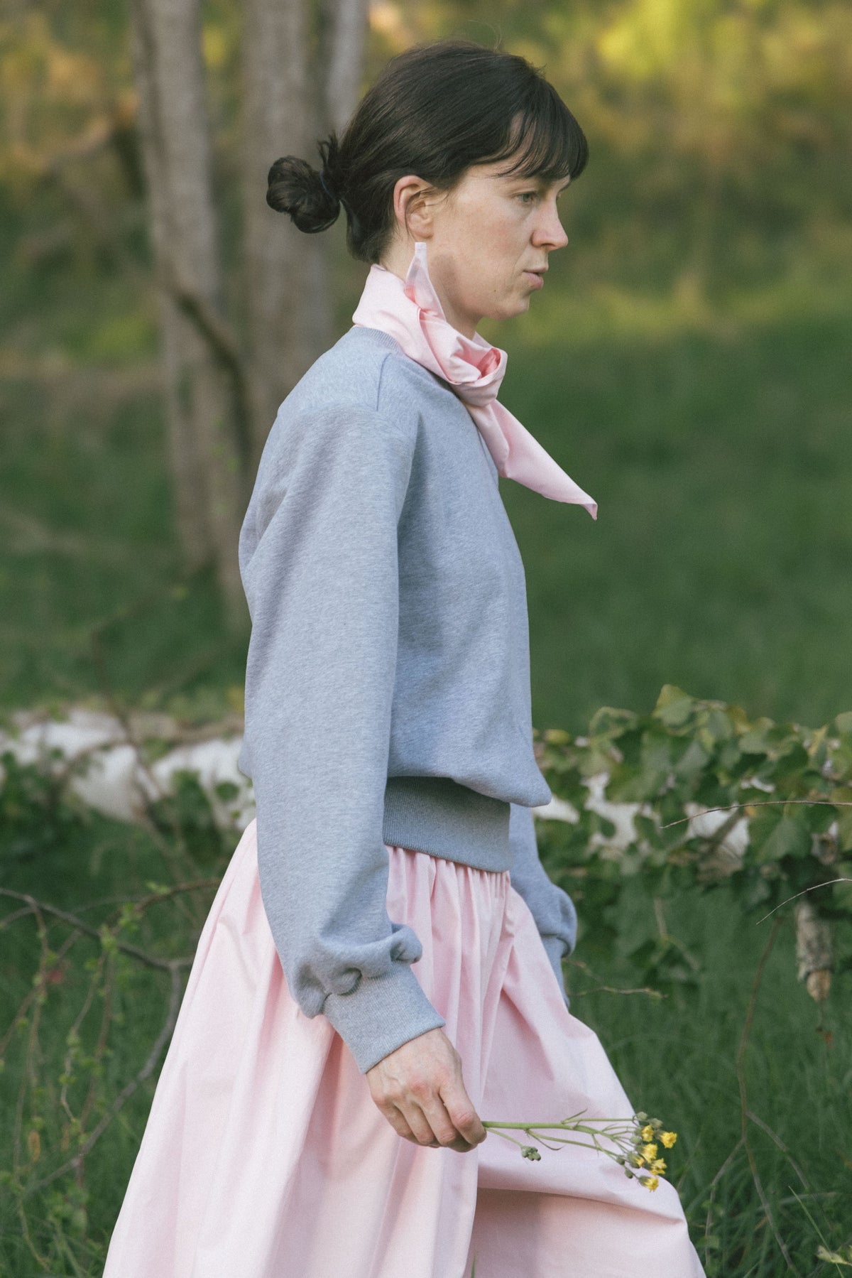 Side view of a model walking outdoors in the light gray Paulette Madeleine Top with pale pink trousers and a pink neck scarf, carrying a small bunch of flowers and showing the loose sleeve gathered at the cuff.