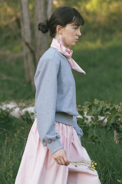 Side view of a model walking outdoors in the light gray Paulette Madeleine Top with pale pink trousers and a pink neck scarf, carrying a small bunch of flowers and showing the loose sleeve gathered at the cuff.