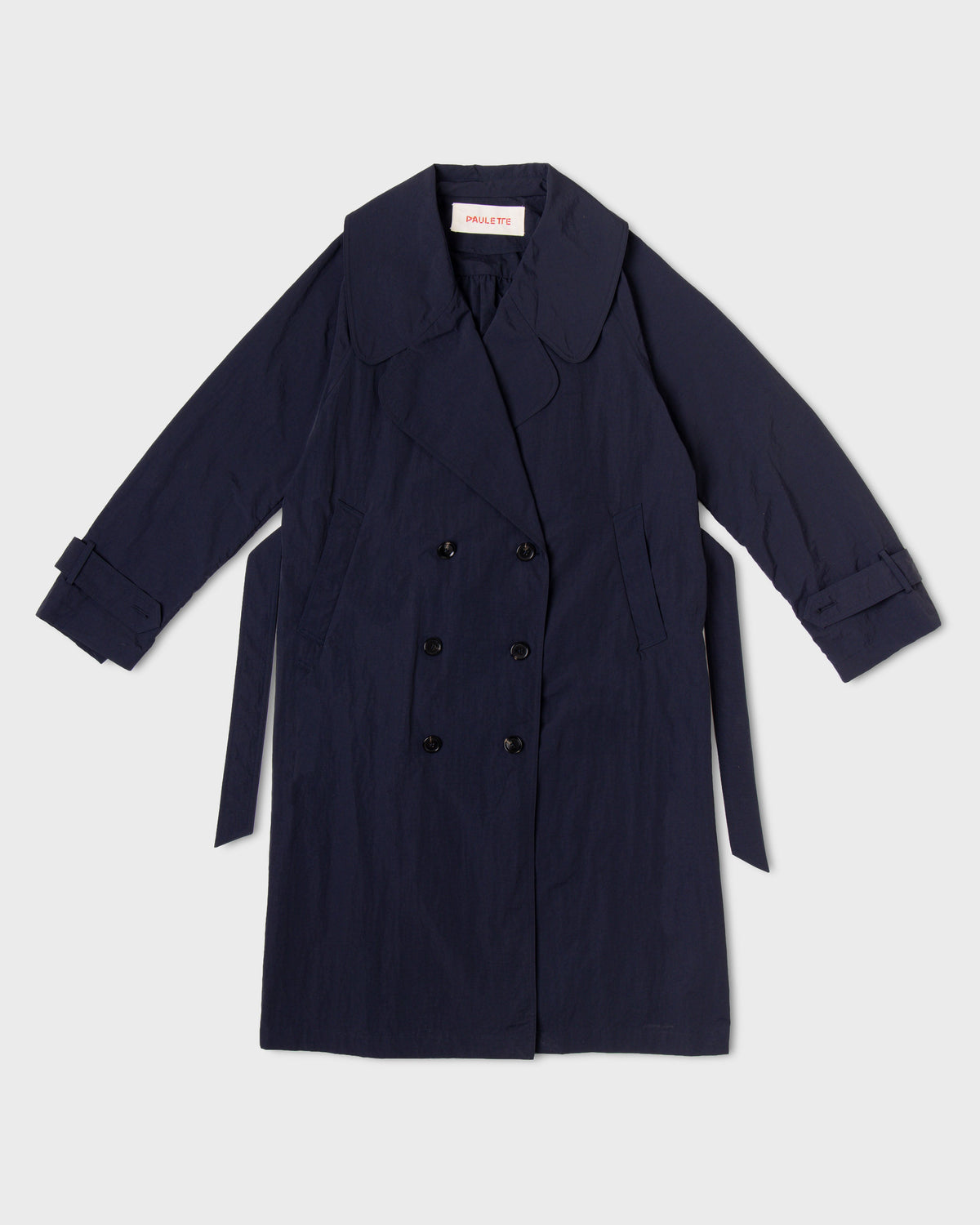 Dark navy Paulette Louise Trench Coat displayed against a light background, showing a broad rounded collar, oversized overlapping lapels, six dark buttons in two rows, slanted pockets, cuff straps, and a matching belt hanging at the sides.