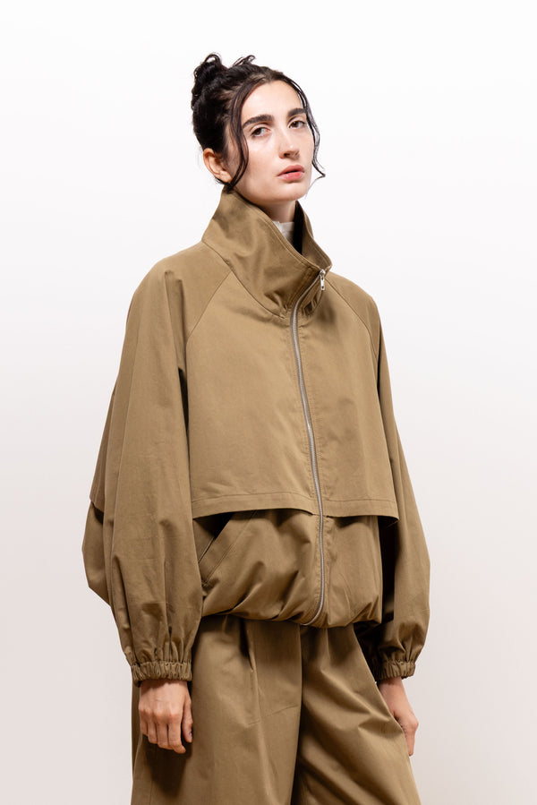 Three-quarter view of a model wearing the khaki-brown Paulette Lee Bomber Jacket zipped up, showing a tall stand collar, dropped shoulders, full sleeves, gathered cuffs and hem, and a wide overlapping panel across the front.