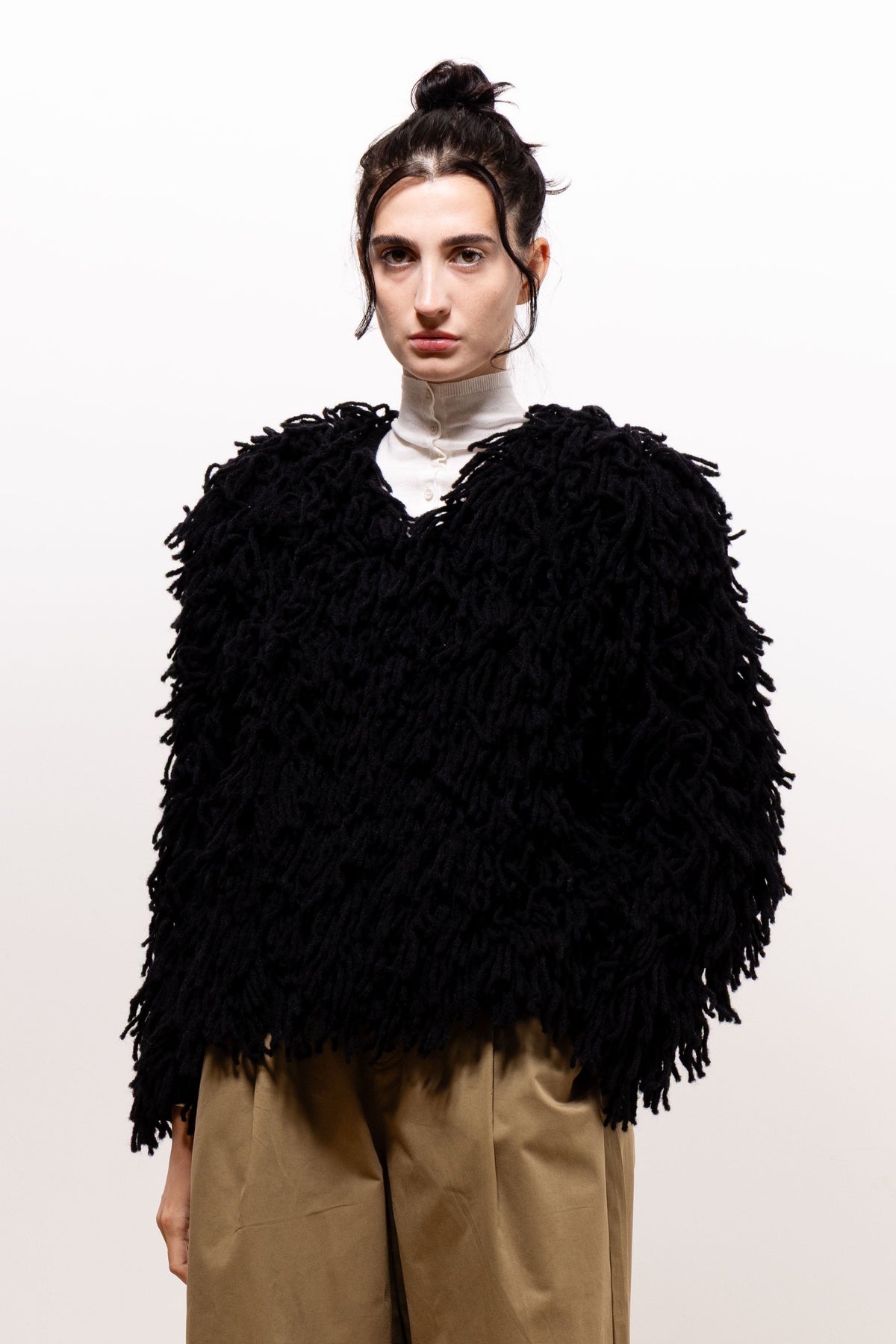 Slightly angled front view of a model wearing the black Paulette Saint-Jean-de-Luz Cardigan, showing its broad shoulders, full sleeves, and long textured strands over khaki-brown trousers.