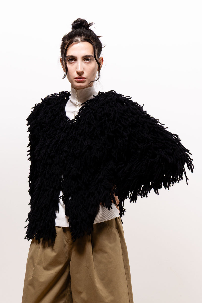 Front view of a model wearing the black Paulette Saint-Jean-de-Luz Oversize Scarf across her shoulders, with one arm extended to spread the textured fabric over a white top and khaki-brown trousers.