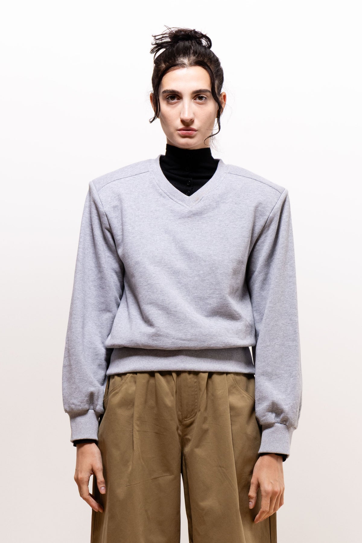 Front view of a model wearing the light gray Paulette Madeleine Top over a black high-neck layer with khaki-brown trousers, showing its shallow V-neck, defined shoulder seams, full long sleeves, and wide ribbed waistband.