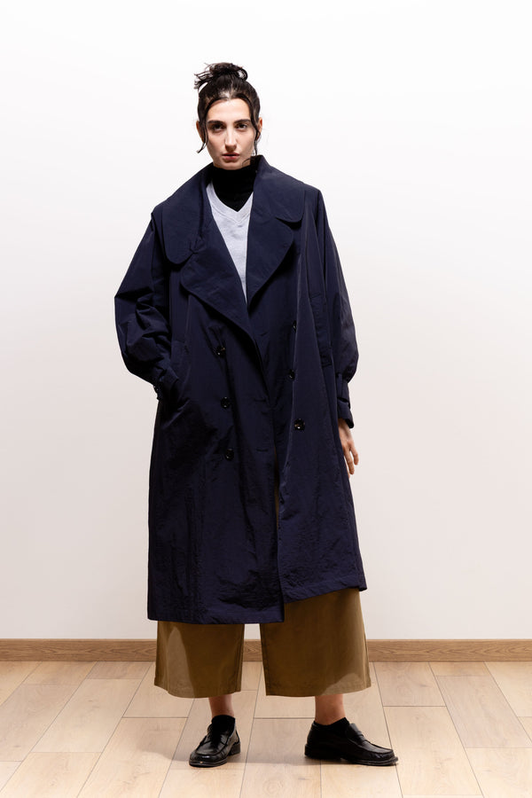 Model wearing the dark navy Paulette Louise Trench Coat unbelted with one hand in a pocket, showing its loose knee-length silhouette and broad lapels over layered tops and khaki-brown trousers.