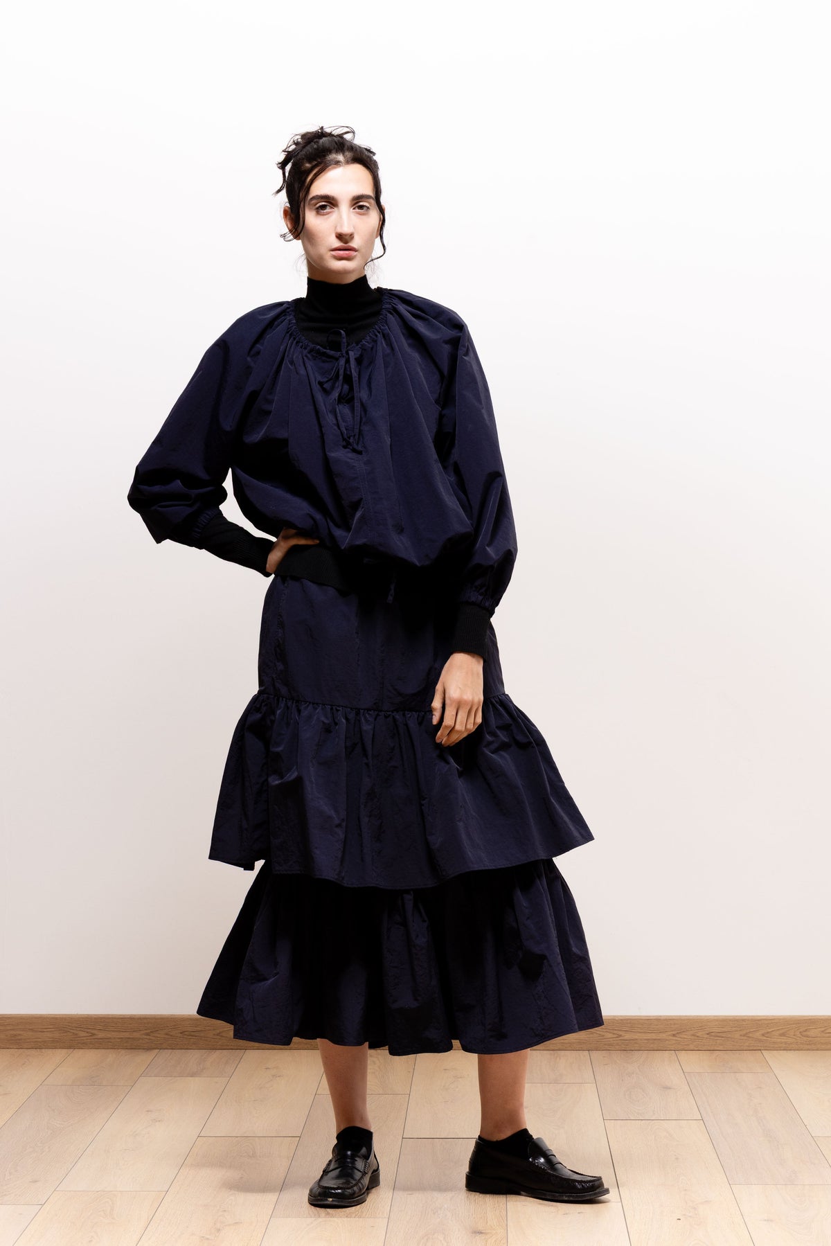 Model wearing the dark navy Paulette Georgia Top with a matching tiered midi skirt and black shoes, posing with one hand on her hip to show its billowing sleeves and gathered waist.