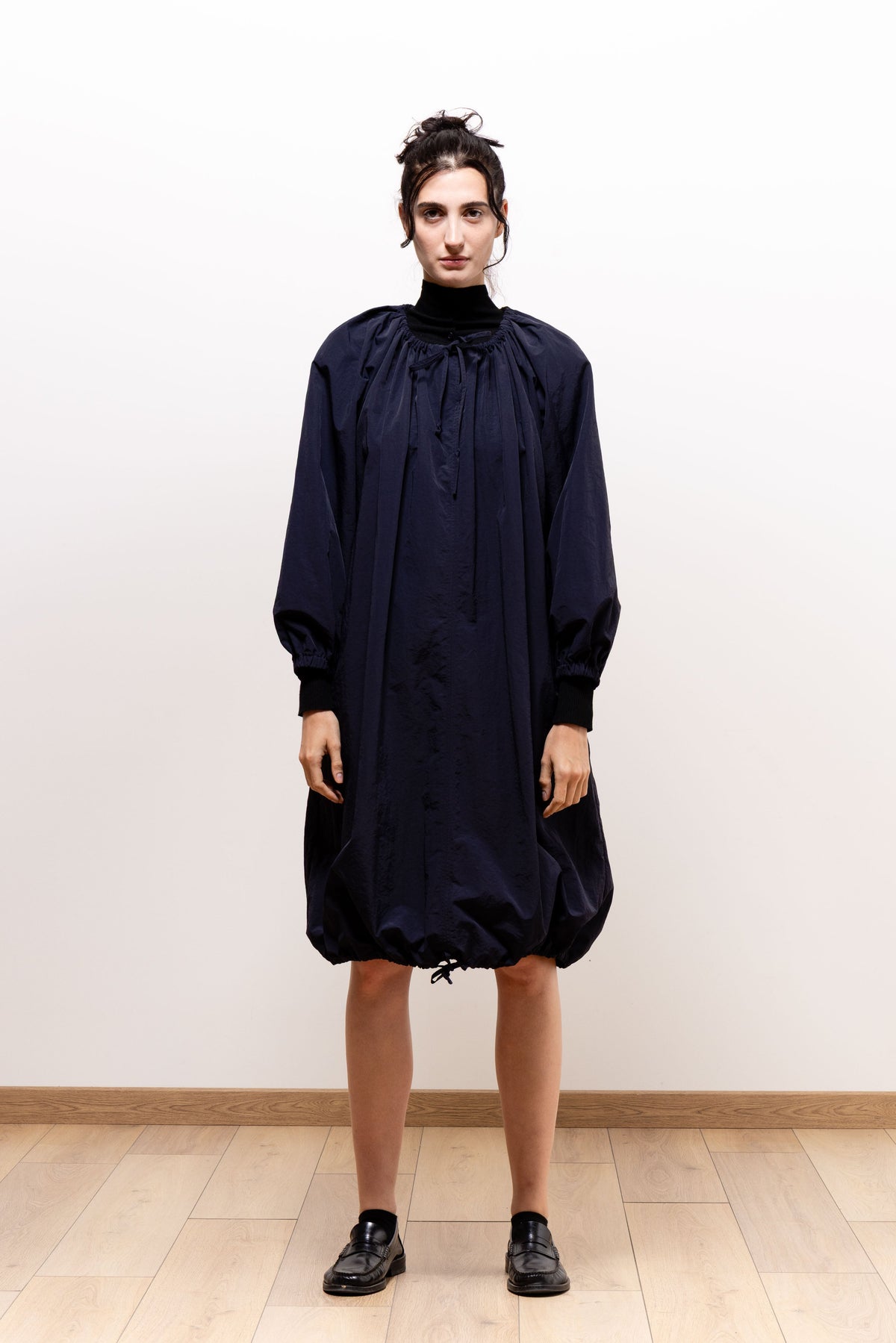 Full-length front view of a model wearing the dark navy Paulette Georgia Dress with black shoes, showing its high neckline, gathered upper front, loose body, and knee-length bubble hem.