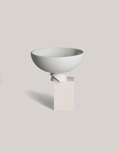 White Tortuga Forma Kaolin Rice Bowl elevated on a rectangular cream display block, showing its smooth rounded body, open interior and sculptural foot formed from overlapping curved pieces against a light grey background.