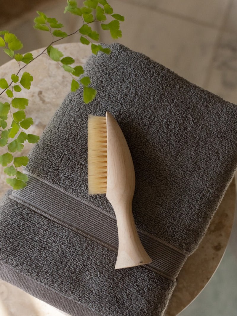 Close-up of a folded charcoal gray towel on a stone surface, with a wooden bath brush resting across its woven border and green foliage nearby.
