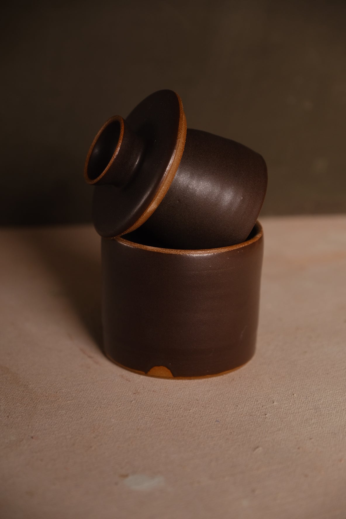 Dark chocolate-brown Settle Ceramics Butterbell on a tan tabletop, with its cup-shaped lid tilted inside the cylindrical base, showing a round knob and contrasting ochre edges.