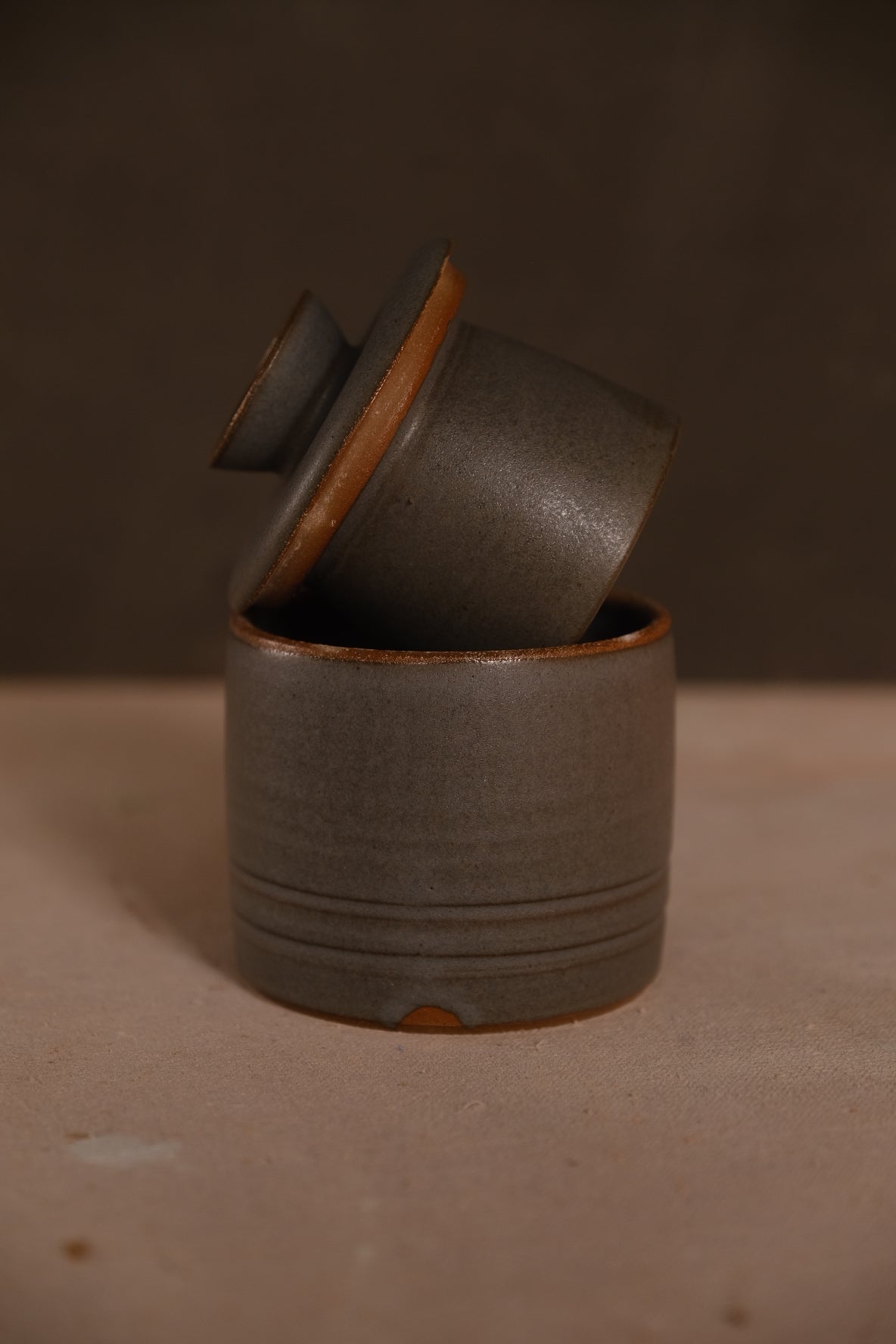 Charcoal-grey Settle Ceramics Butterbell with fine horizontal ridges around the base, an ochre-edged lid and a dark round knob, displayed partly open on a tan surface.