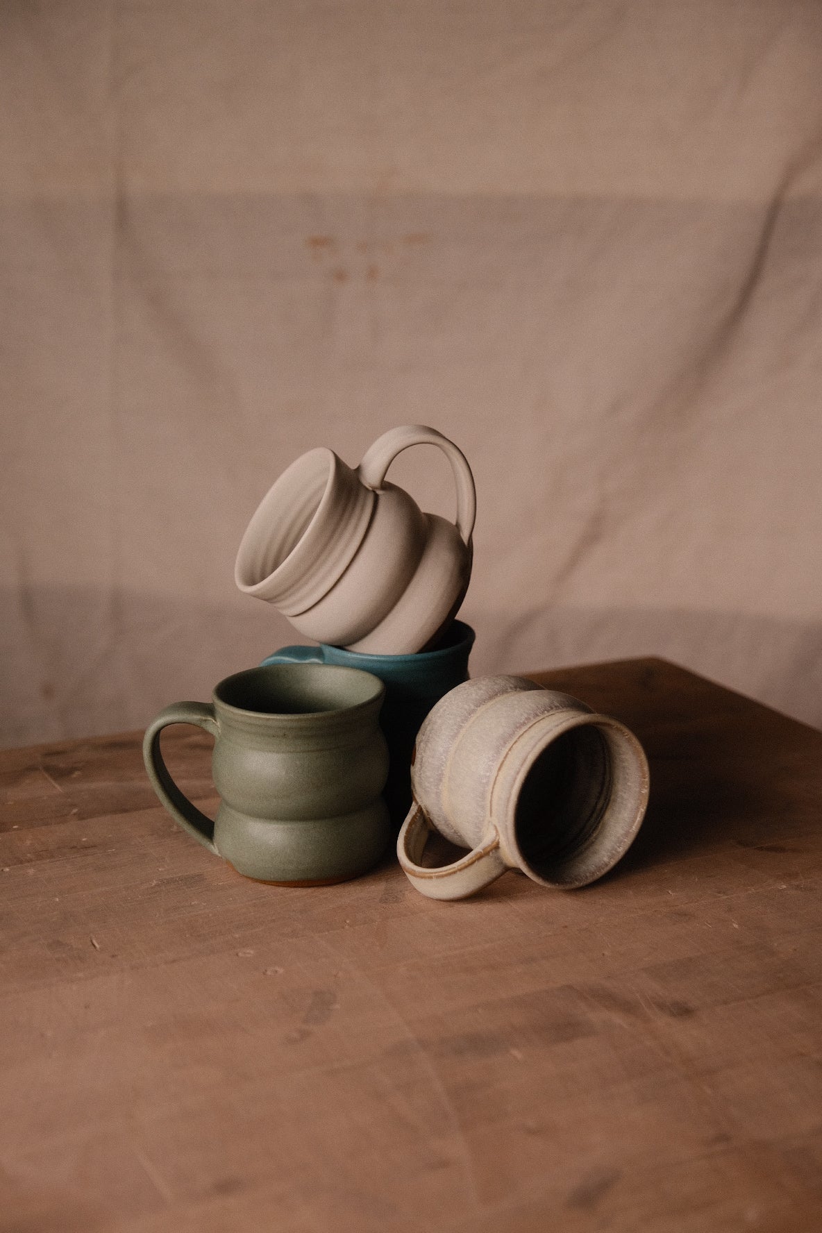 Settle Ceramics The Wave Mugs in cream, green and dark blue arranged against a beige fabric backdrop, with upright, tilted and sideways mugs showing their curved handles, rounded profiles and contrasting interiors.