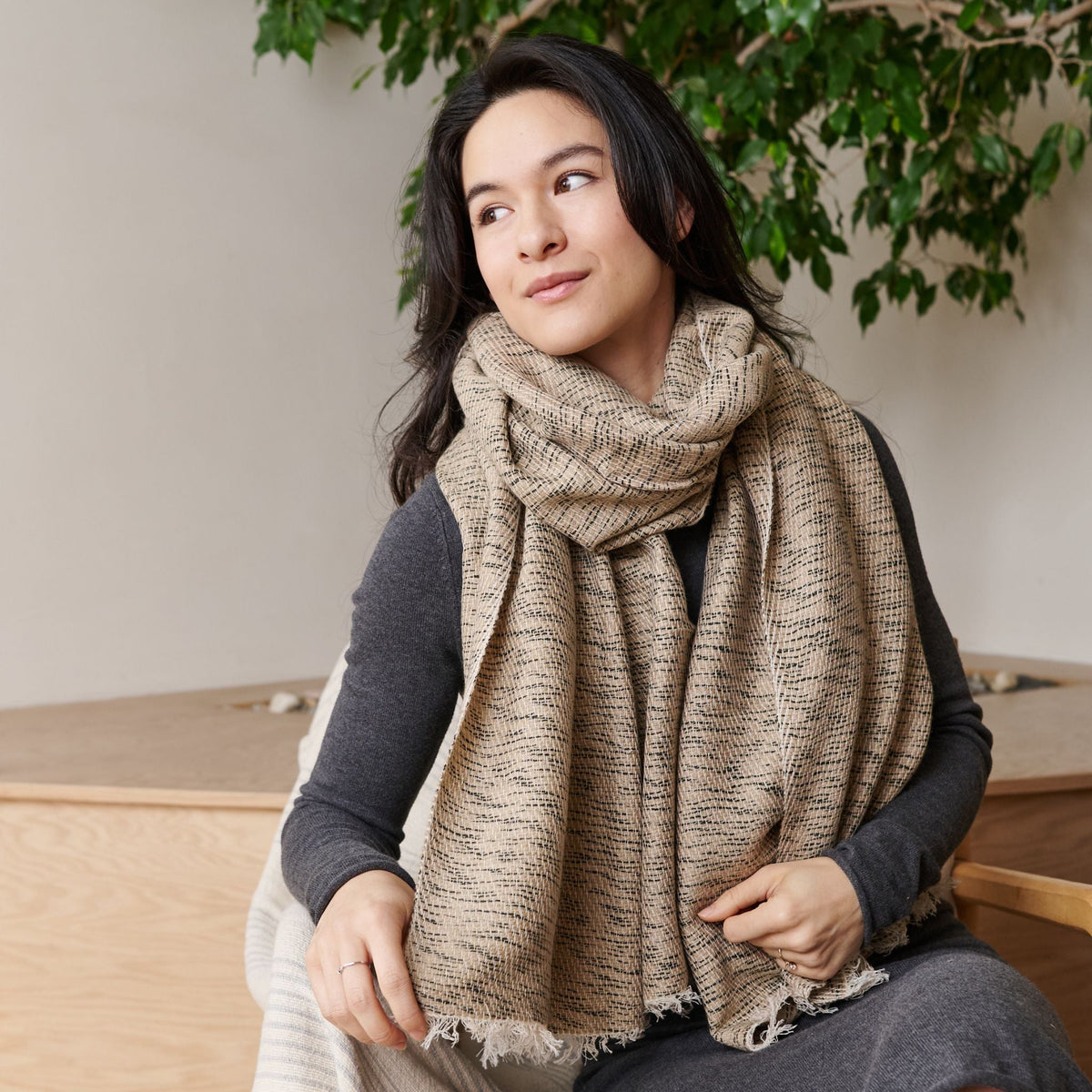 Woman wearing the oversized neutral beige Forest Night scarf, showing its dark woven flecks and softly fringed edges.