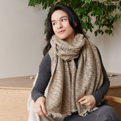 Woman wearing the oversized neutral beige Forest Night scarf, showing its dark woven flecks and softly fringed edges.