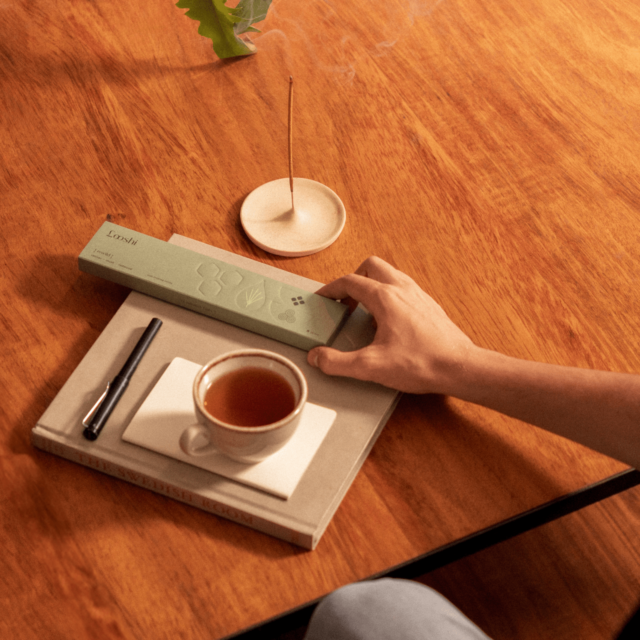 Overhead view of a person holding a Looshi Rewild incense box at a wooden table with tea, a book and a burning incense stick
